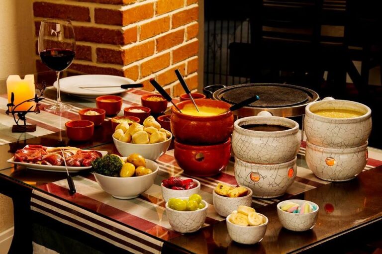 Depois de um dia conhecendo parques, paisagens e atrações da Serra Gaúcha, a programação não precisa terminar. Gramado à noite oferece restaurantes, experiências gastronômicas, noites temáticas e passeios pelo centro que ajudam a aproveitar ainda mais a viagem. Há opções para diferentes perfis e momentos do roteiro. Enquanto alguns visitantes procuram o tradicional fondue, outros preferem conhecer a cultura regional por meio da gastronomia, da música e das noites típicas. Por isso, vale reservar pelo menos algumas noites da viagem para descobrir Gramado depois que anoitece. O que fazer à noite em Gramado? A programação noturna de Gramado está bastante ligada à gastronomia e ao entretenimento. Uma viagem de poucos dias, por exemplo, pode combinar uma noite dedicada ao fondue, outra para uma experiência cultural típica e uma terceira mais livre para caminhar pelo centro e escolher um restaurante. Além disso, a programação muda ao longo do ano. Eventos, temporadas especiais e períodos como o Natal Luz acrescentam novas possibilidades ao destino. Portanto, antes de viajar, é interessante pensar nas noites como parte do roteiro, e não apenas decidir o que fazer depois que os passeios diurnos terminarem. Fondue: um clássico das noites em Gramado Poucas experiências gastronômicas são tão associadas a Gramado quanto o fondue. O clima da Serra Gaúcha ajudou a transformar a sequência de fondue em uma das escolhas mais tradicionais para o jantar. Normalmente, a experiência reúne diferentes etapas, que podem incluir queijos, carnes e chocolate. É uma alternativa especialmente procurada por casais, famílias e grupos que desejam reservar uma noite para uma refeição mais demorada. A JE Hoffmann Turismo trabalha com a Noite Suíça, permitindo incluir essa tradição gastronômica na programação da viagem. Como os restaurantes podem ter bastante movimento em períodos de alta temporada, organizar essa experiência com antecedência pode facilitar o planejamento.